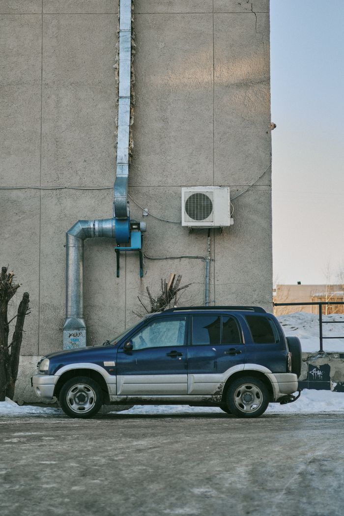 services-03 A blue SUV parked beside a building with industrial piping and an AC unit on a snowy day.